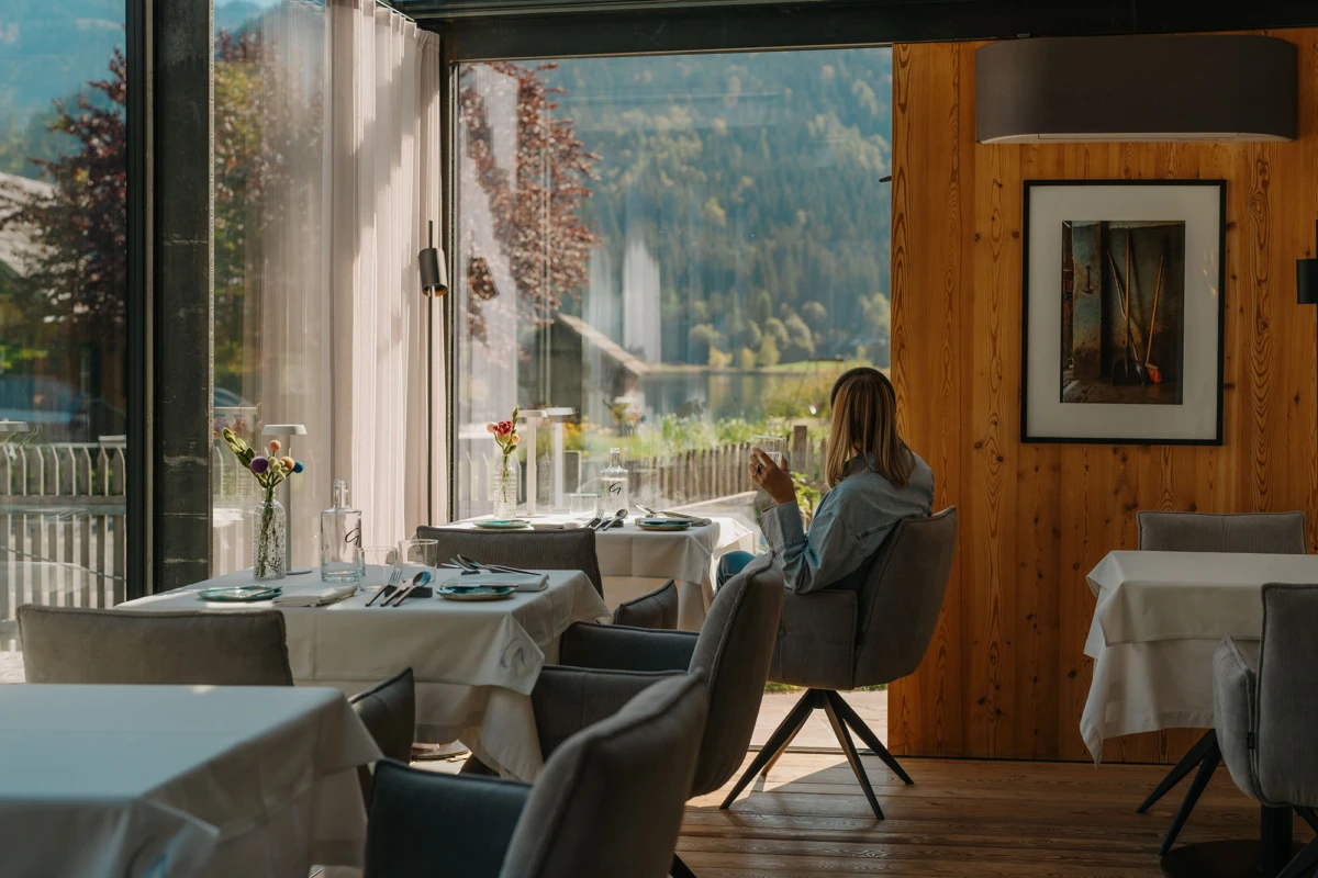Person sitzt im Restaurant am Fenster, schaut auf Smartphone; Holzwand, Blumenvasen, weiße Tische, Aussicht auf Berge und See.