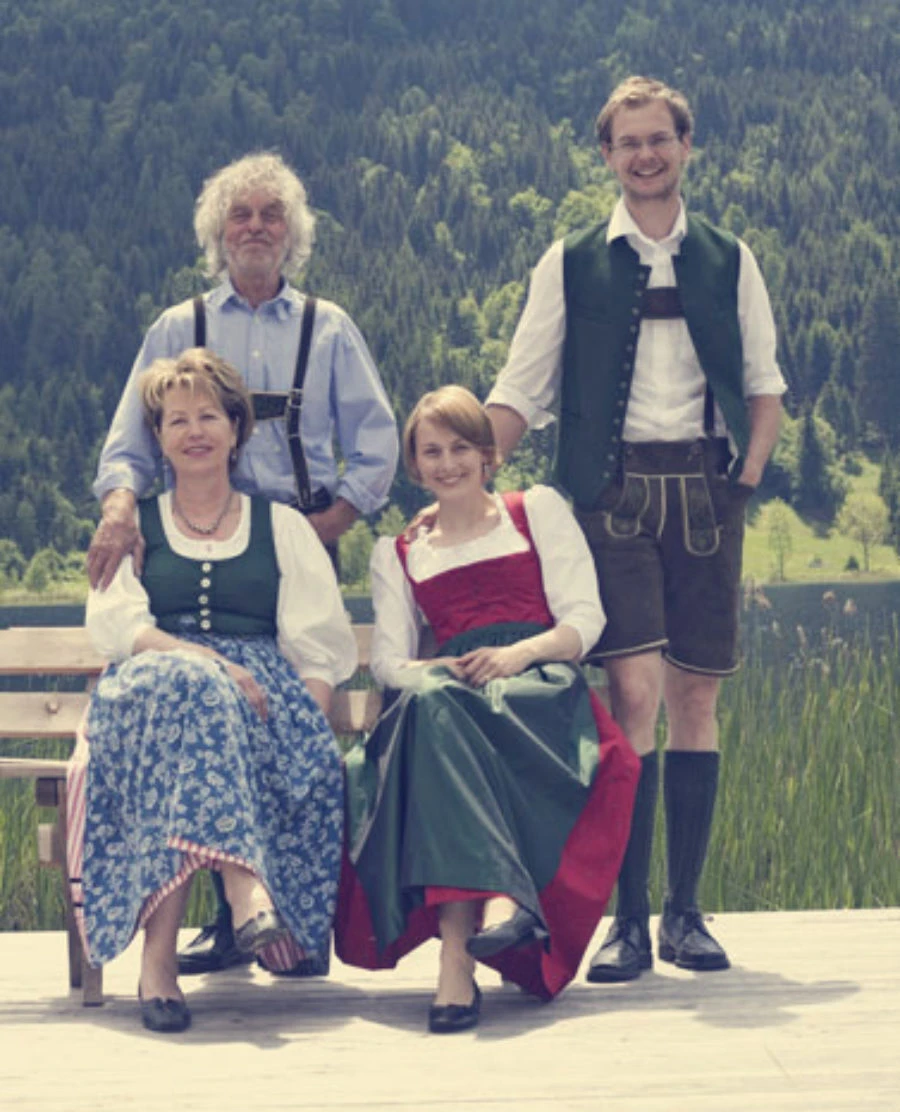 Vier Personen in Dirndl und Lederhosen posieren auf einem Holzsteg am See, Berglandschaft im Hintergrund.