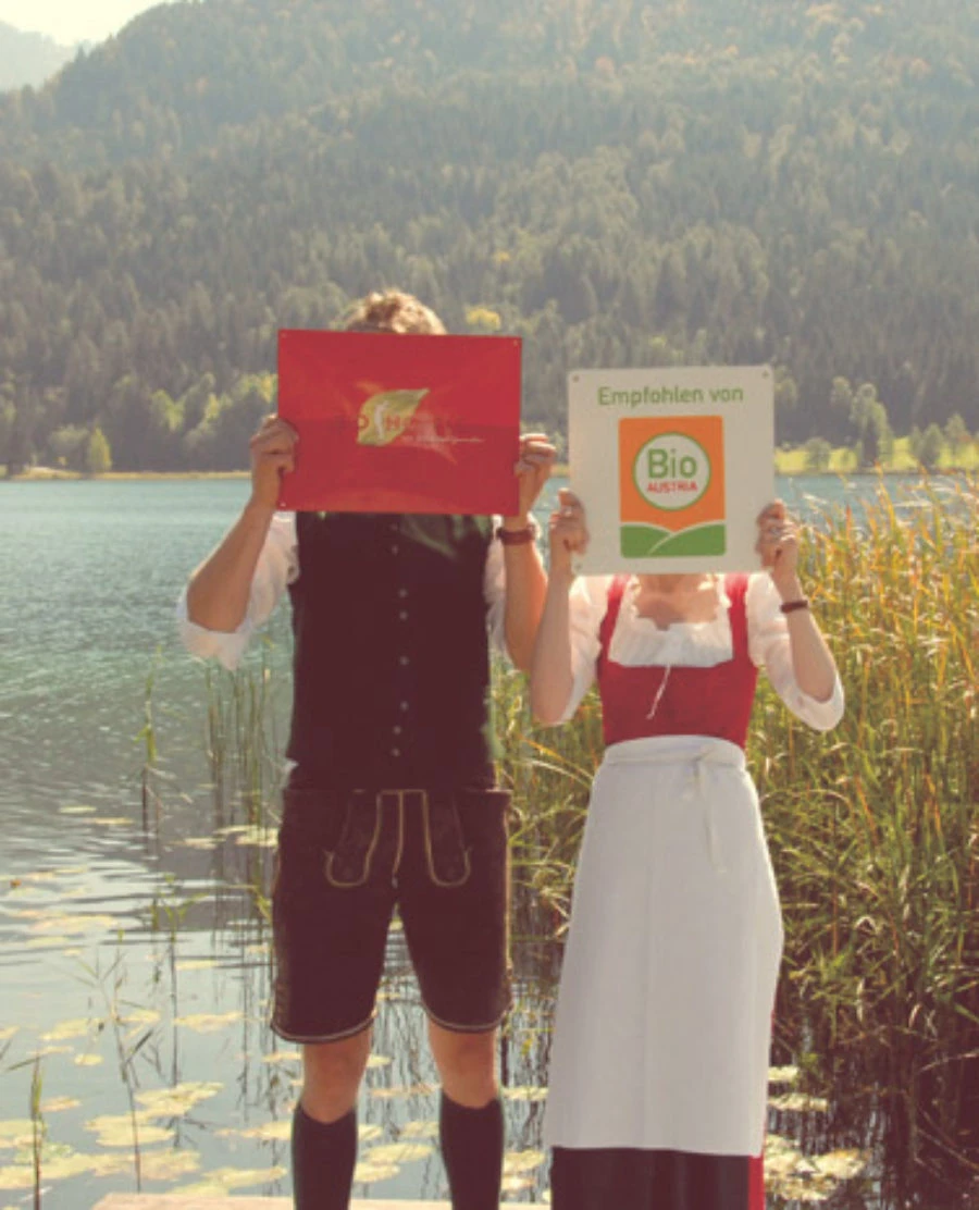Zwei Personen in Trachten stehen am Seeufer und halten Schilder vor dem Gesicht; rotes Schild links, Schild mit Bio Austria rechts.