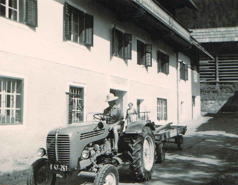 Historischer Traktor mit Fahrer in Hut am Steuer, zieht Anhänger vor weißem Gebäude mit Fensterläden.