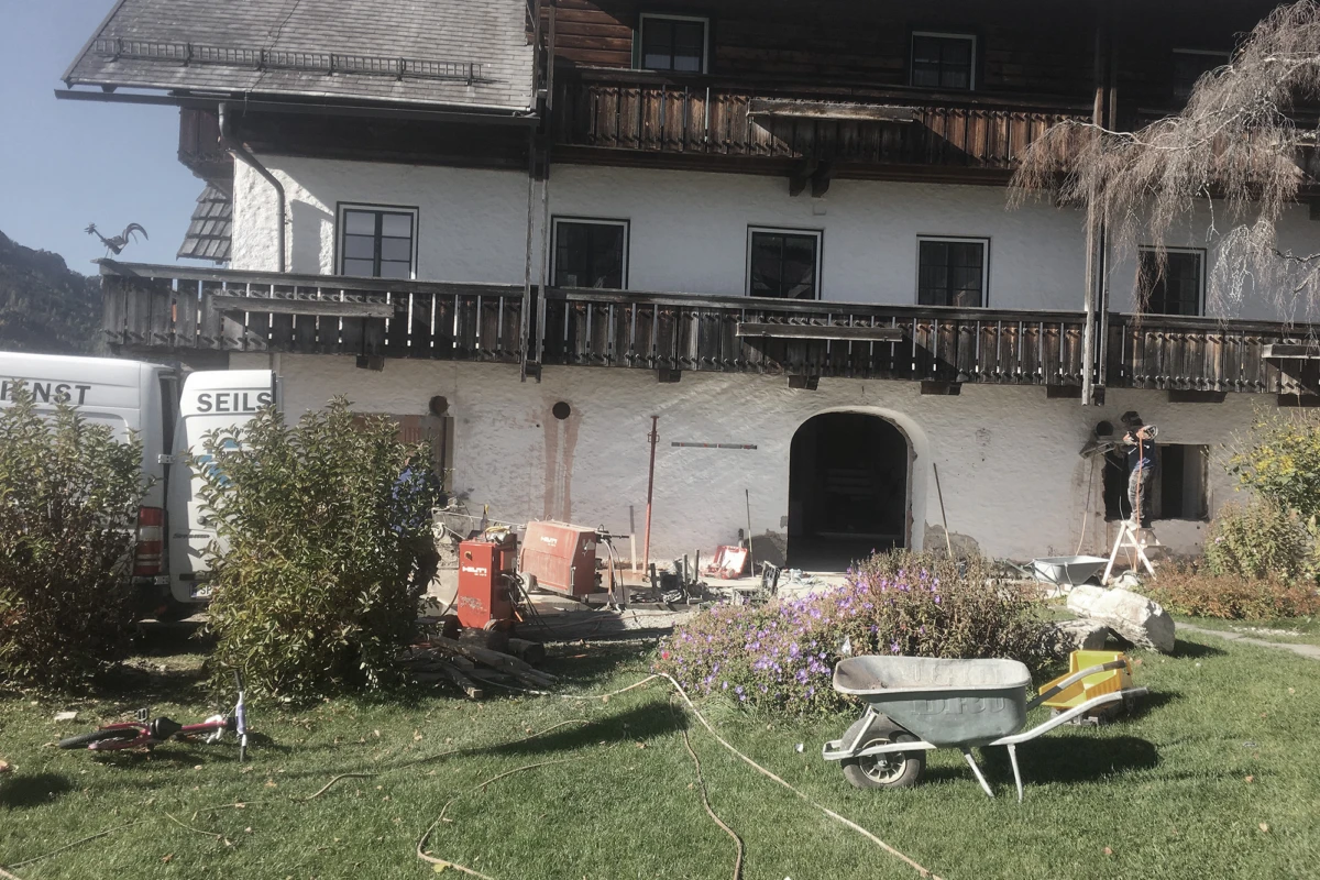 Chaletartiges Haus mit weißer Fassade und dunklen Holzbalkonen; Renovierungsarbeiten im Vorgarten, Werkzeuge, Schubkarre und Leiter.