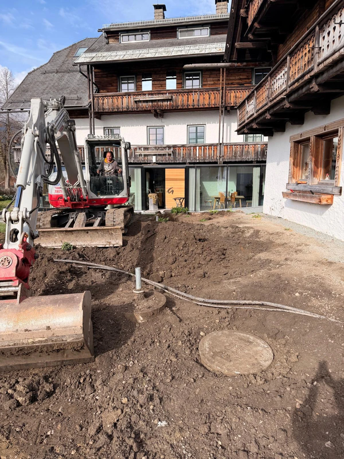 Minibagger mit Fahrer gräbt Erdreich vor einem mehrstöckigen Chalet-Haus mit Holzbalkonen.