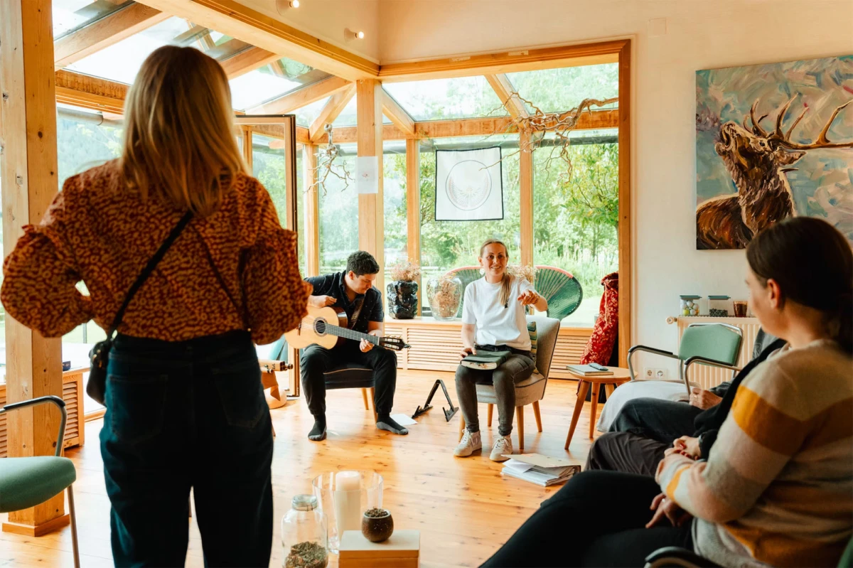 Gitarrist spielt Akustikgitarre, umgeben von weiteren Personen in hellem Holzstudio mit großen Fenstern.