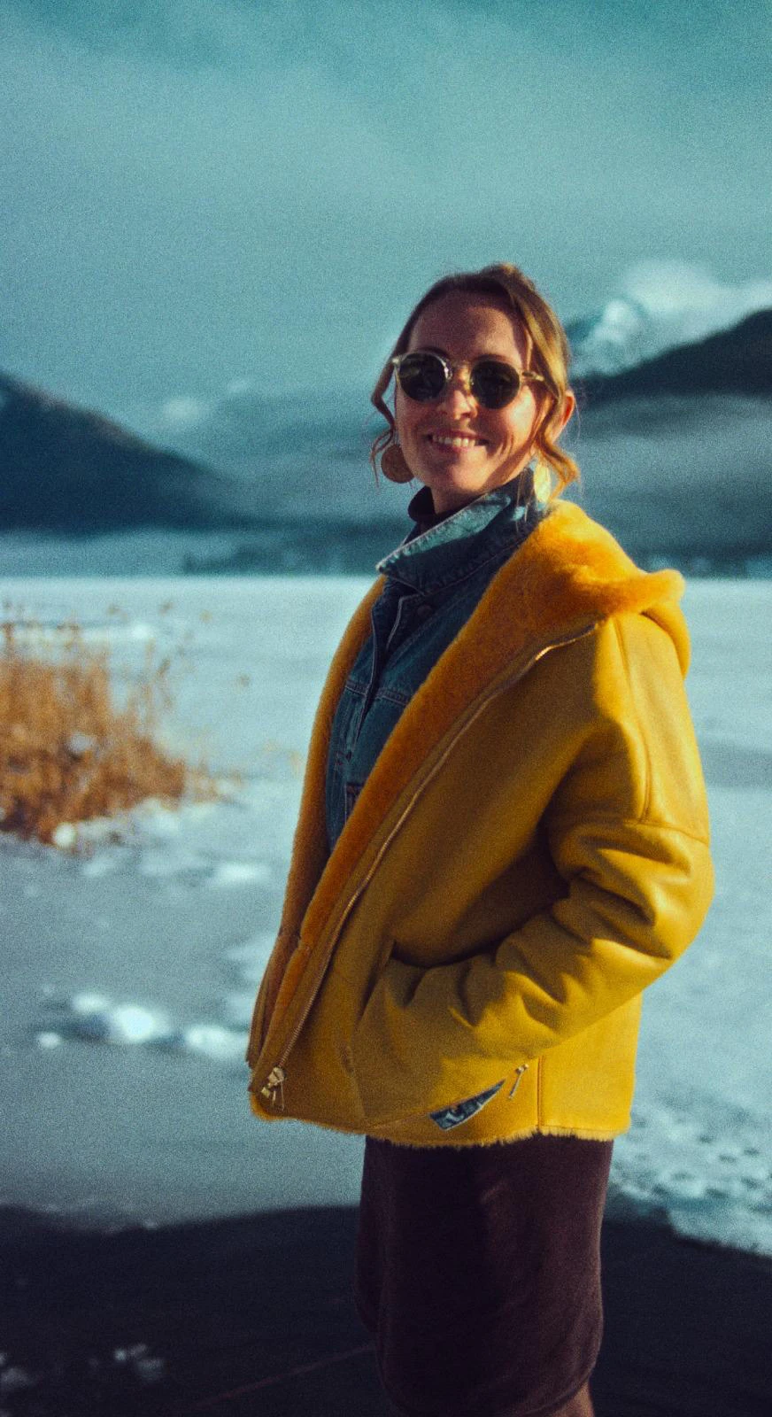Person in senfgelben Mantel mit Jeansjacke und Sonnenbrille steht lächelnd in Winterlandschaft mit Schnee und Bergen