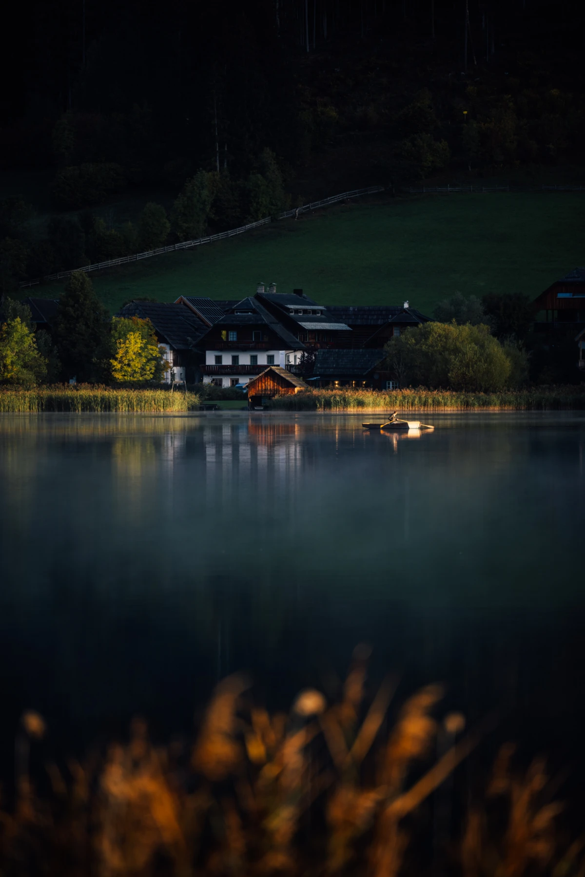 Dorf am Seeufer im Abendlicht; ruhiges Wasser, kleines Boot treibt vor dem Bootshaus aus Holz, von Bergen und Wald umgeben.