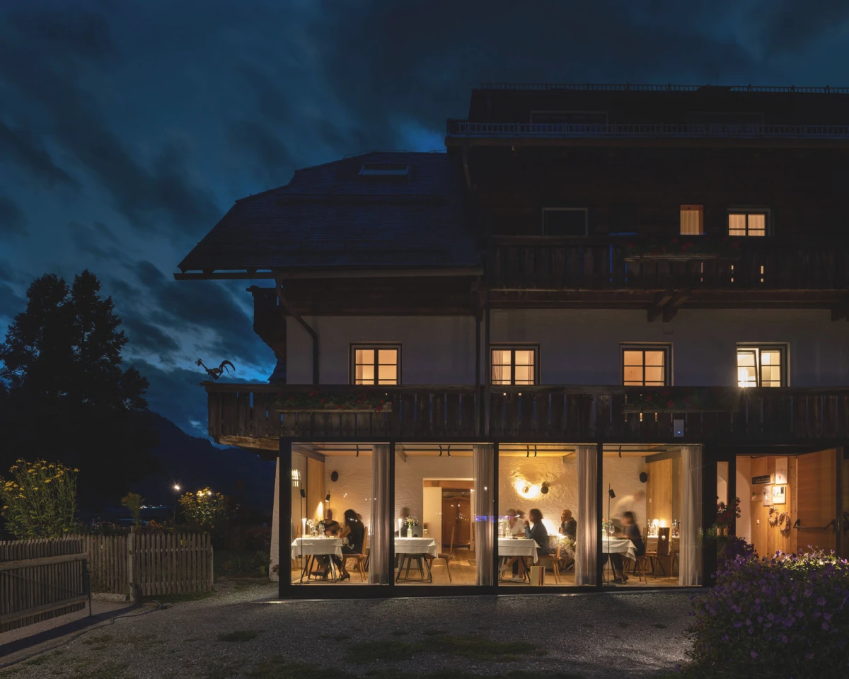 Alpenchalet-Gasthaus mit Glasfront, abends, Gäste speisen im Innenraum.