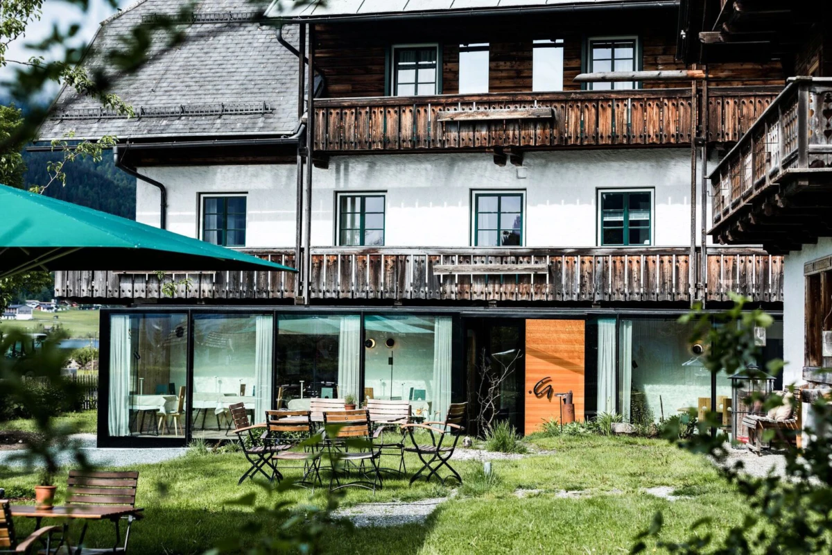 Chaletartiges Hotelgebäude mit Holzbalustraden und großer Glasfront; Außenbereich mit Holztischen und Stühlen auf grünem Rasen.