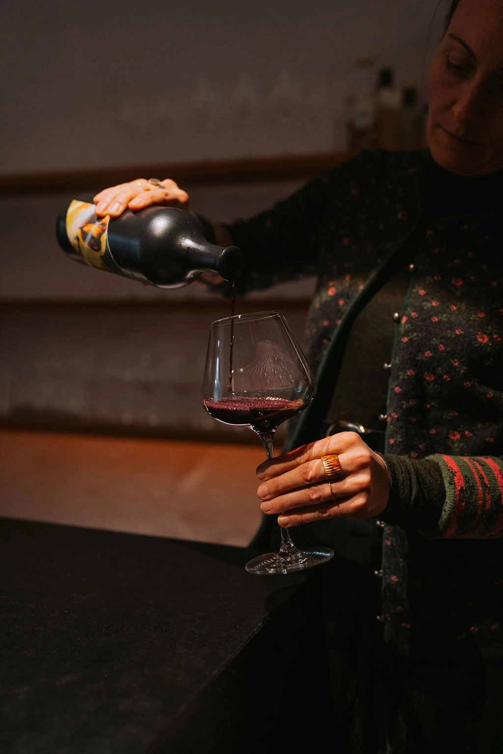 Person gießt Rotwein aus Flasche in Weinglas am dunklen Bartresen.