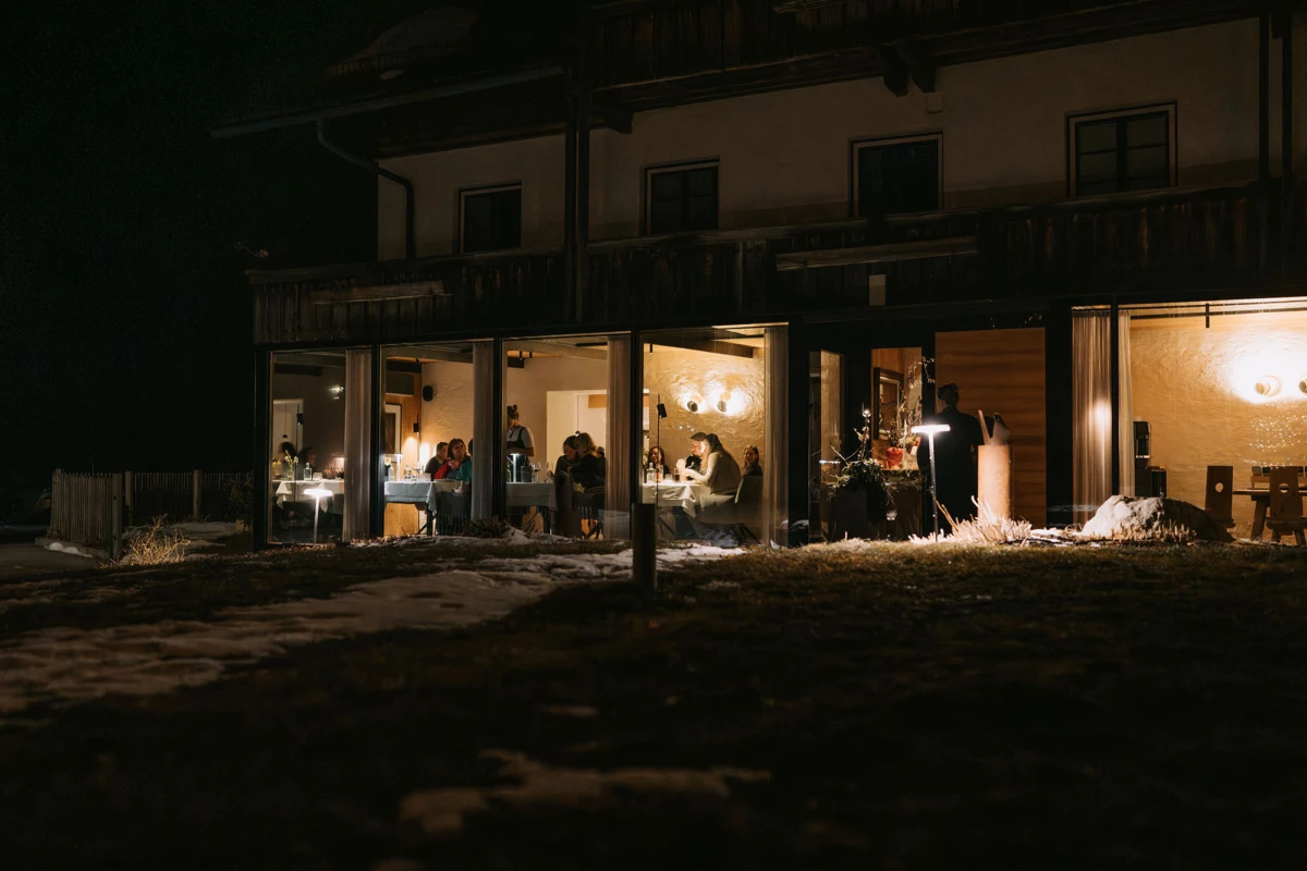 Nächtliche Dinner-Szene in Glas-Wintergarten eines Hauses; mehrere Personen essen an Tischen, warmes Licht, Schnee draußen.