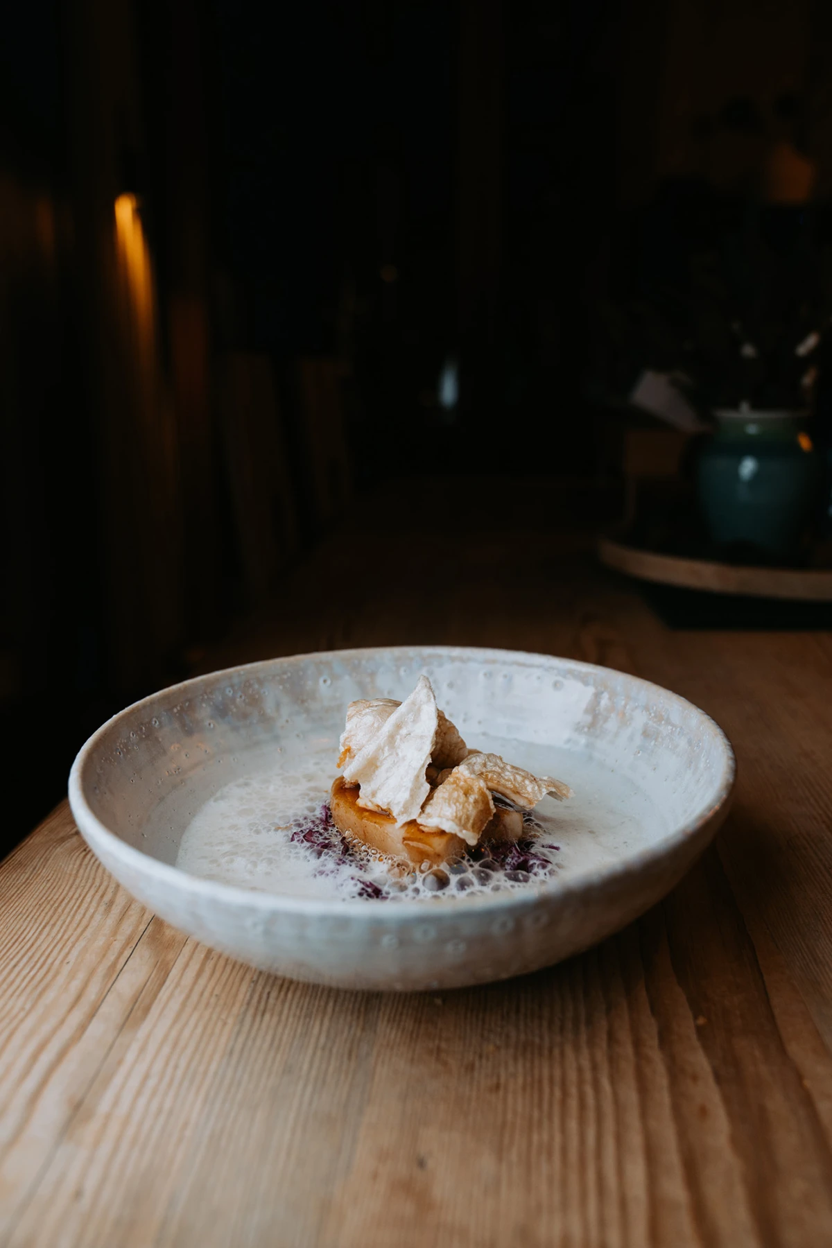 Graue Keramikschale mit cremigem Dessert, garniert mit knusprigen Stückchen, auf holzfarbenem Tisch vor dunklem Hintergrund.