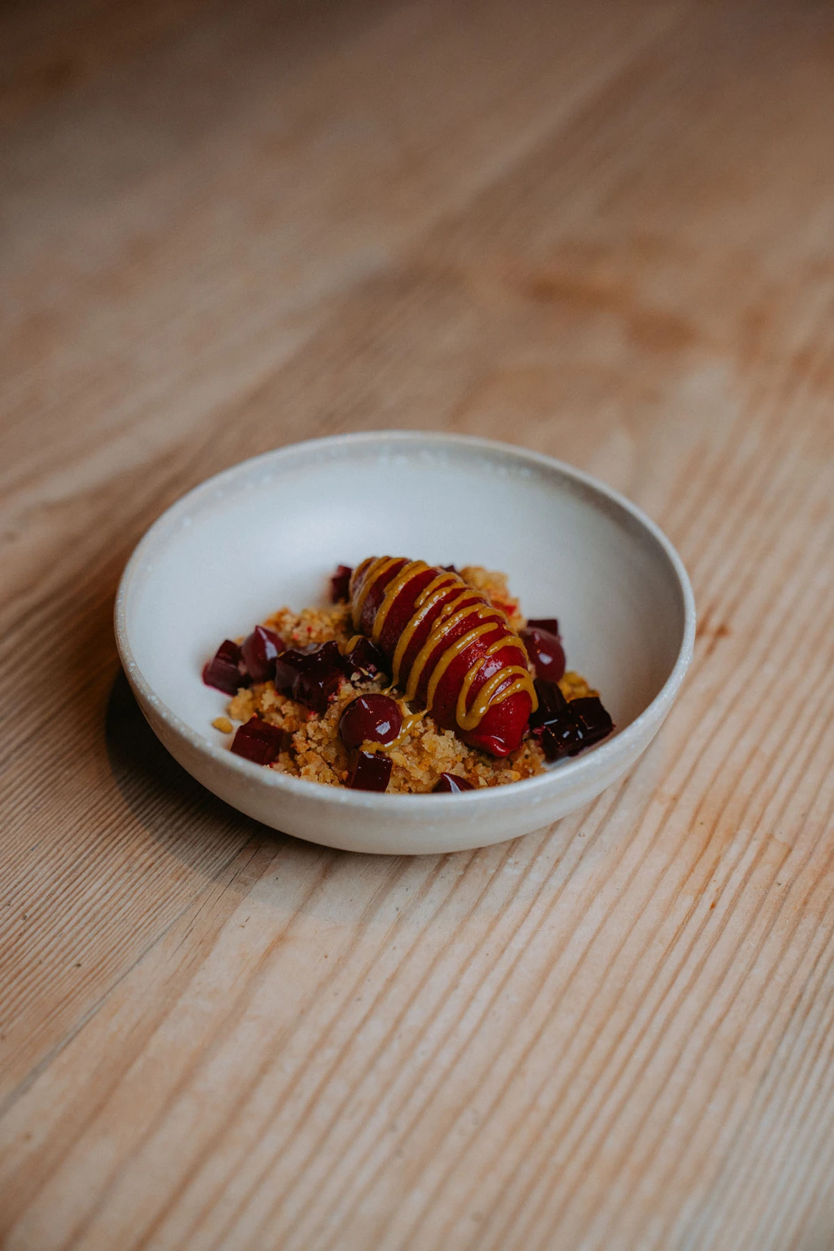 Weiße Schüssel mit Frucht-Crumble und roten Beeren; längliche gelbe Glasur zieht darüber, auf Holztisch.