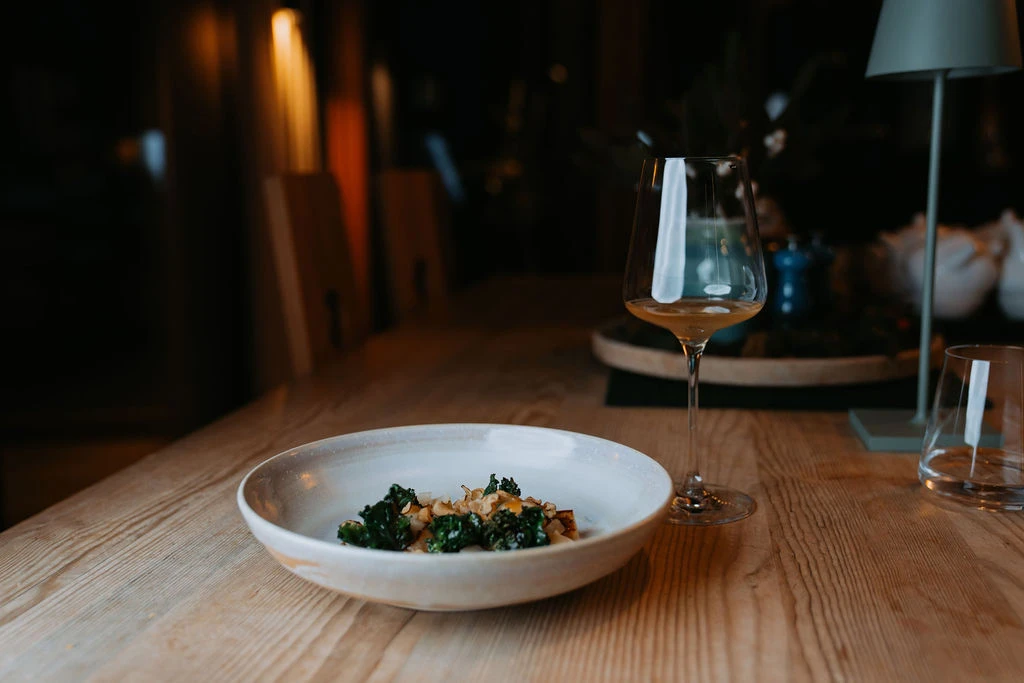 Schüssel mit Grünkohl und Croutons auf Holztisch; daneben Weinglas, gedämpftes Restaurantlicht im Hintergrund.