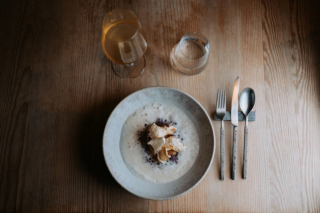 Keramikschale mit cremiger Suppe und Croutons auf Holztisch; daneben Wasser- und Weinglas, Besteck rechts.