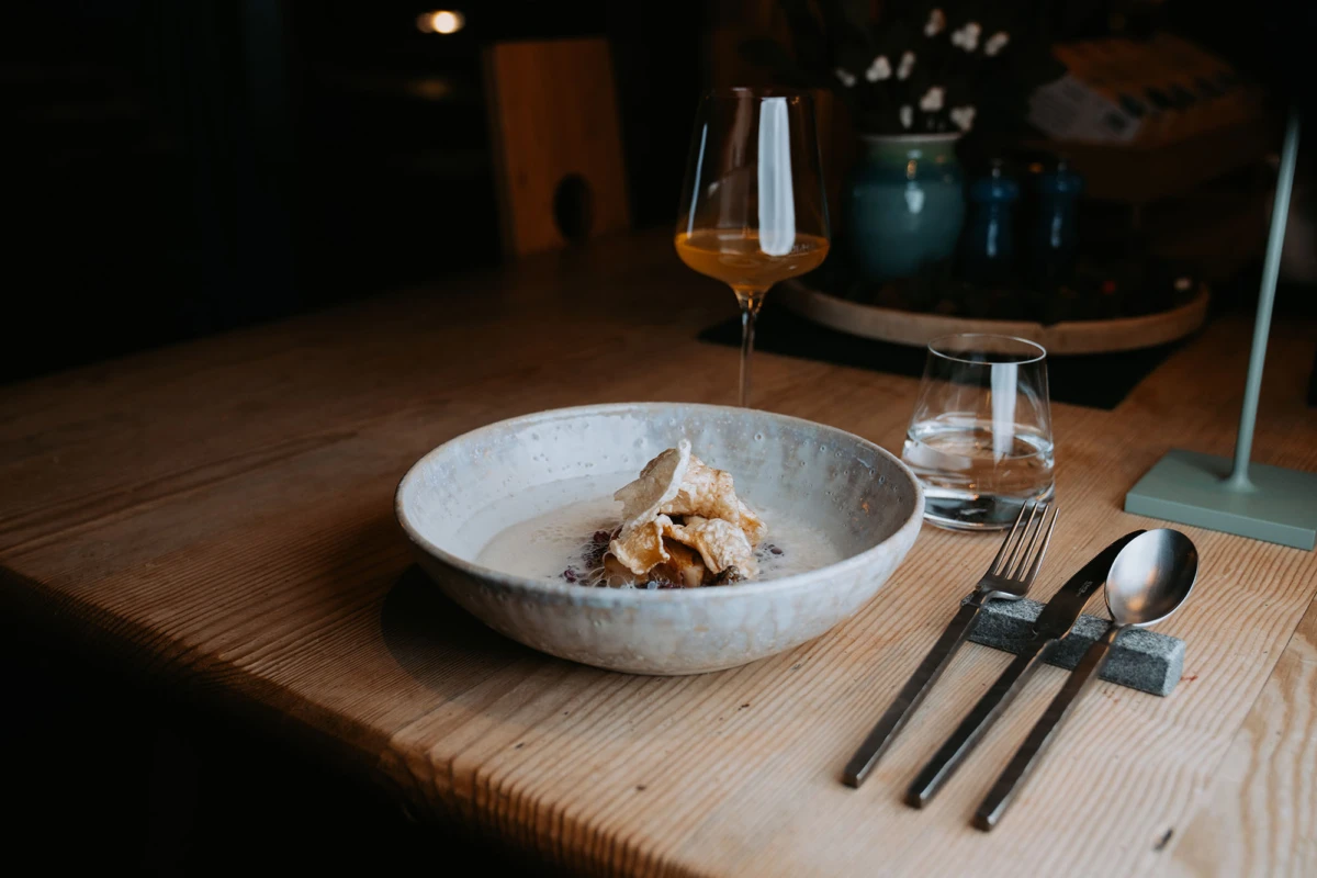 Keramikschale mit Dessert und knusprigen Blätterteigstücken auf Holztisch; Wasser- und Weinglas, Besteck im Restaurant.