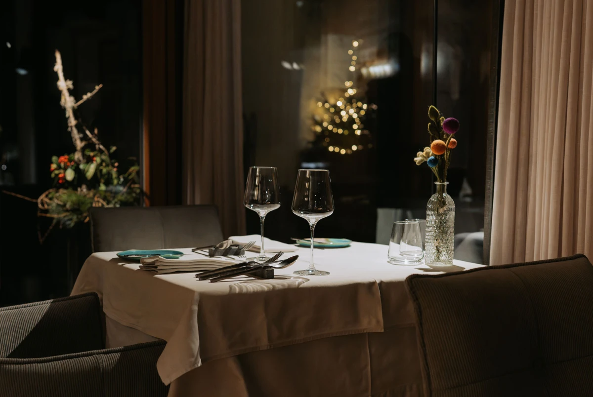 Gedeckter Esstisch im Restaurant: weiße Tischdecke, zwei Weingläser, Besteck, Teller; Vase mit bunten Trockenblumen, Vorhänge im Hintergrund.
