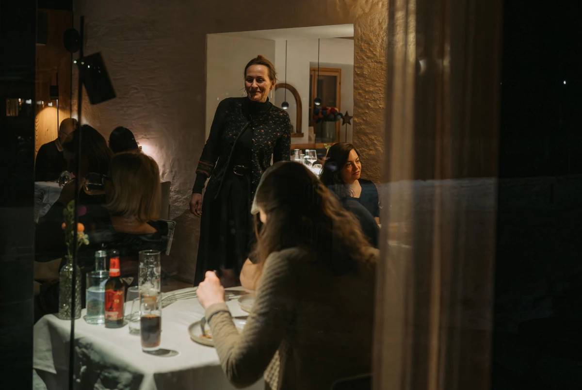 Gastgeberin in dunklem Mantel steht am Kopf eines gedeckten Tisches in einem gemütlichen Restaurant und spricht zu den Gästen.