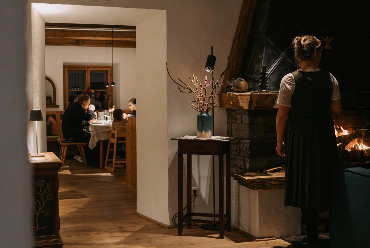Person in traditionellem Kleid steht am Kaminfeuer, im Hintergrund essen Gäste an einem Esstisch in rustikaler Gaststube.