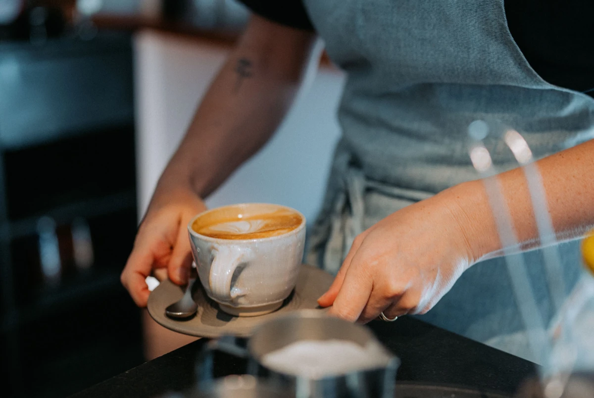 Barista bereitet Latte in Keramikbecher auf Untertasse zu, während eine Hand Löffel hält, in einer Café-Umgebung.