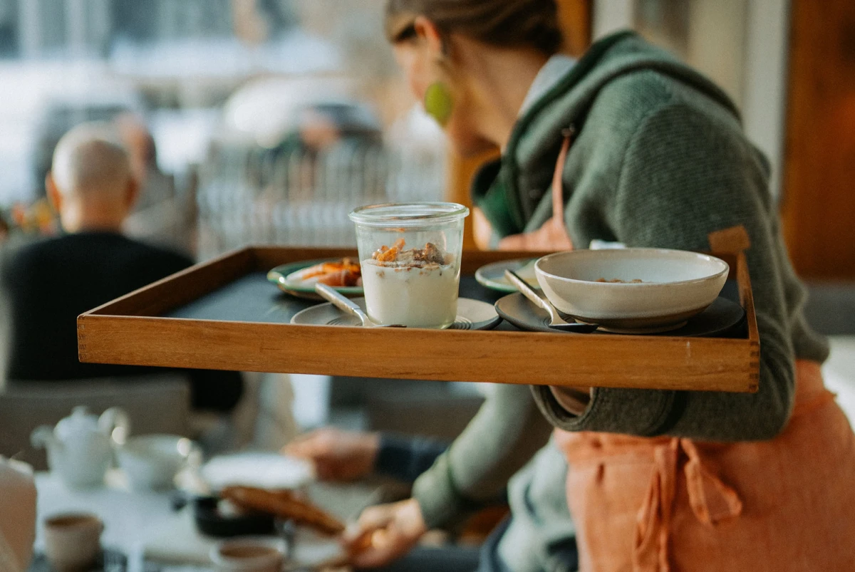 Servicekraft trägt Holztablett mit Joghurtglas, Schüssel und Besteck in einem Café.