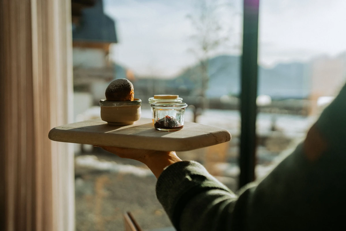 Hand hält Holztablett mit Brötchen in Keramikschale und Marmeladenglas; Fenster mit Berglandschaft im Hintergrund.