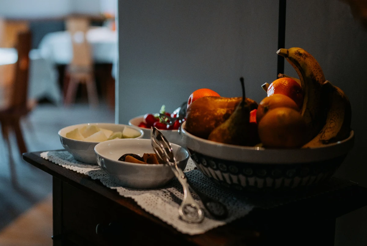 Große Obstschale mit Bananen, Äpfeln und Birnen auf dunklem Sideboard, daneben zwei kleinere Schalen mit Obst auf einer Spitzen-Tischdecke.