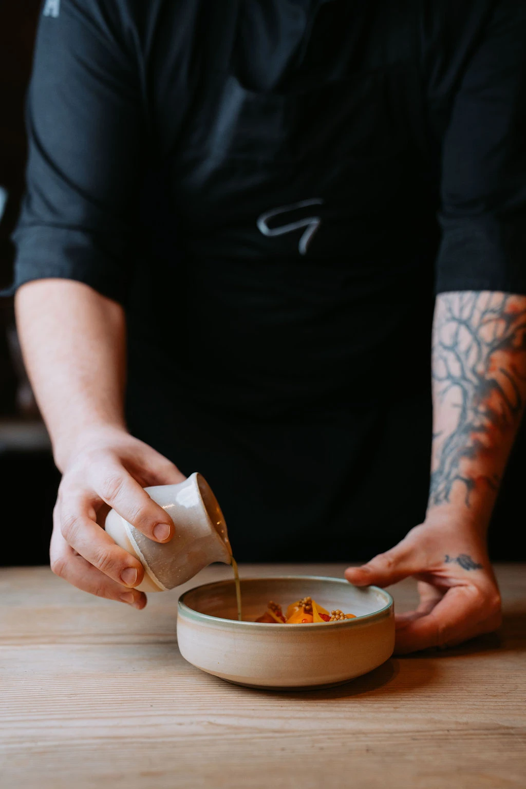 Person mit Unterarmtattoo gießt braune Soße aus kleinem Becher auf Keramikschale mit Snacks auf Holztisch.