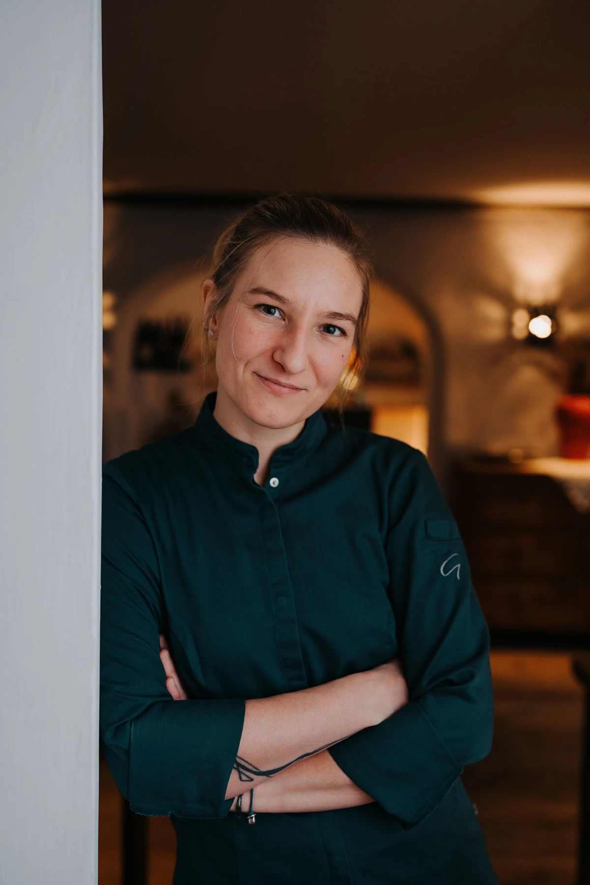 Person in dunkelgrüner Uniform lehnt an weißer Säule, verschränkte Arme, lächelt; warm beleuchteter Innenraum im Hintergrund.