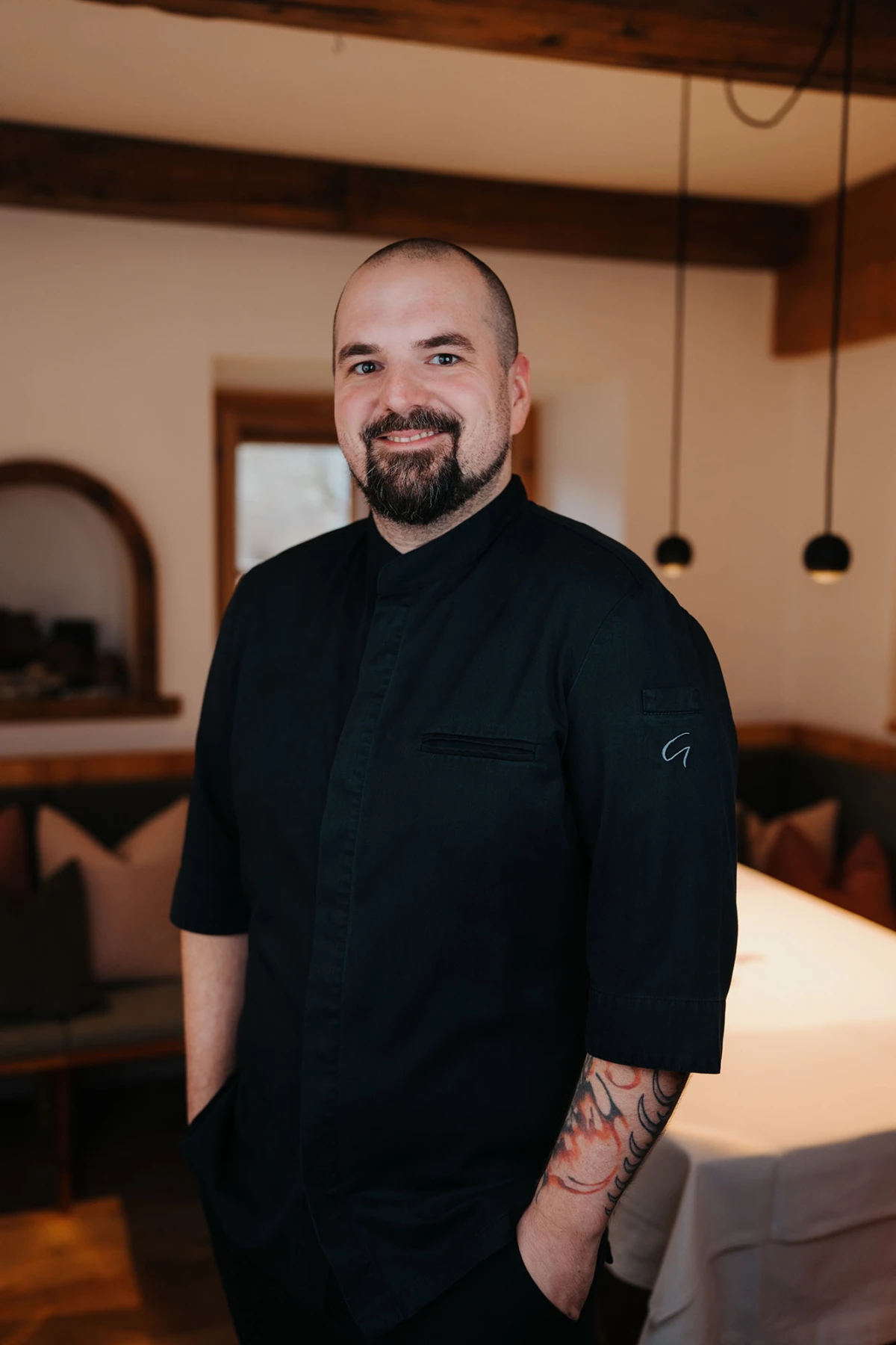 Person in schwarzer Kochjacke steht lächelnd in hellem Restaurant mit Holzbalken und Pendelleuchten.