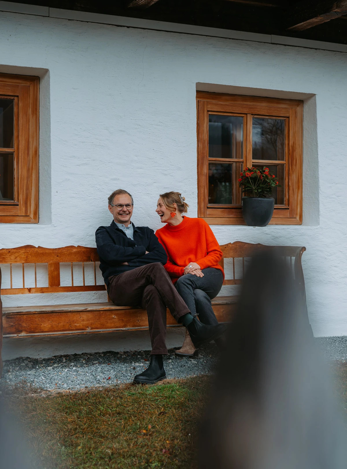 Paar sitzt lächelnd auf einer Holzbank vor einer weißen Hauswand mit Fenster, Blumenkübel mit roten Blumen daneben.
