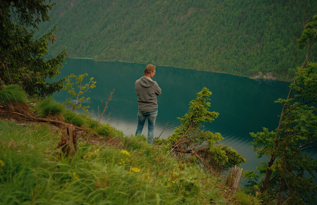 Person mit Hoodie und Jeans steht auf grünem Hang am türkisblauen Bergsee und blickt auf die Waldlandschaft.