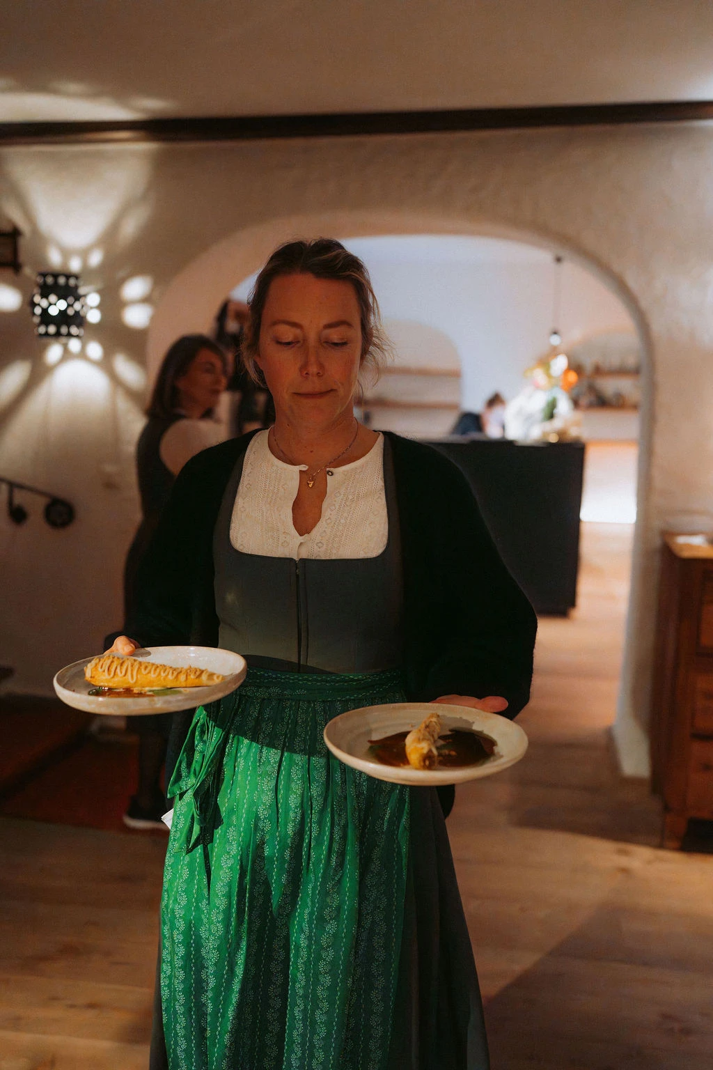 Person in Dirndl serviert zwei Teller Gerichte in gemütlichem Gastraum mit Bögen im Hintergrund.