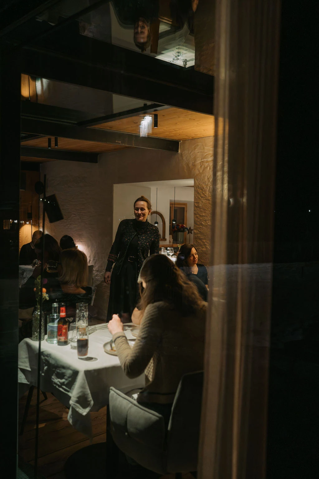 Stehende Person in dunklem Kleid steht an einem gedeckten Tisch in einem gemütlichen Restaurant mit weiteren Gästen und Kerzenlicht.