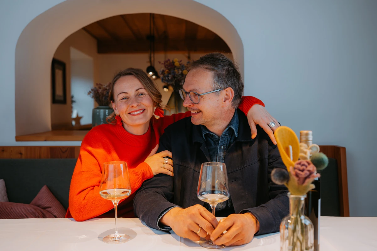 Paar sitzt an einem Tisch in einem Restaurant; Frau in rotem Pullover legt Arm um Mann, beide lächeln; zwei Weingläser, Vase mit Blumen.