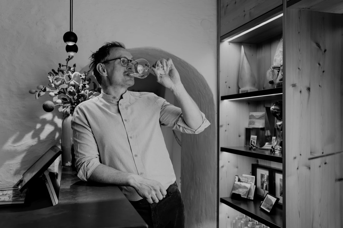 Person mit Brille trinkt Wein aus Weinglas an Theke; links Vase mit Blumen, rechts Holzregale mit Bildern.