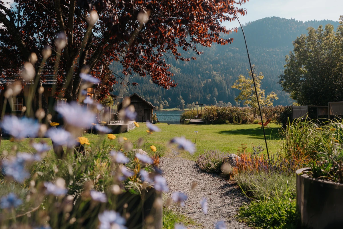 Gartenweg aus Kies führt zu See vor bewaldeten Bergen; bunte Blumen im Vordergrund, Holzhäuschen am Ufer.