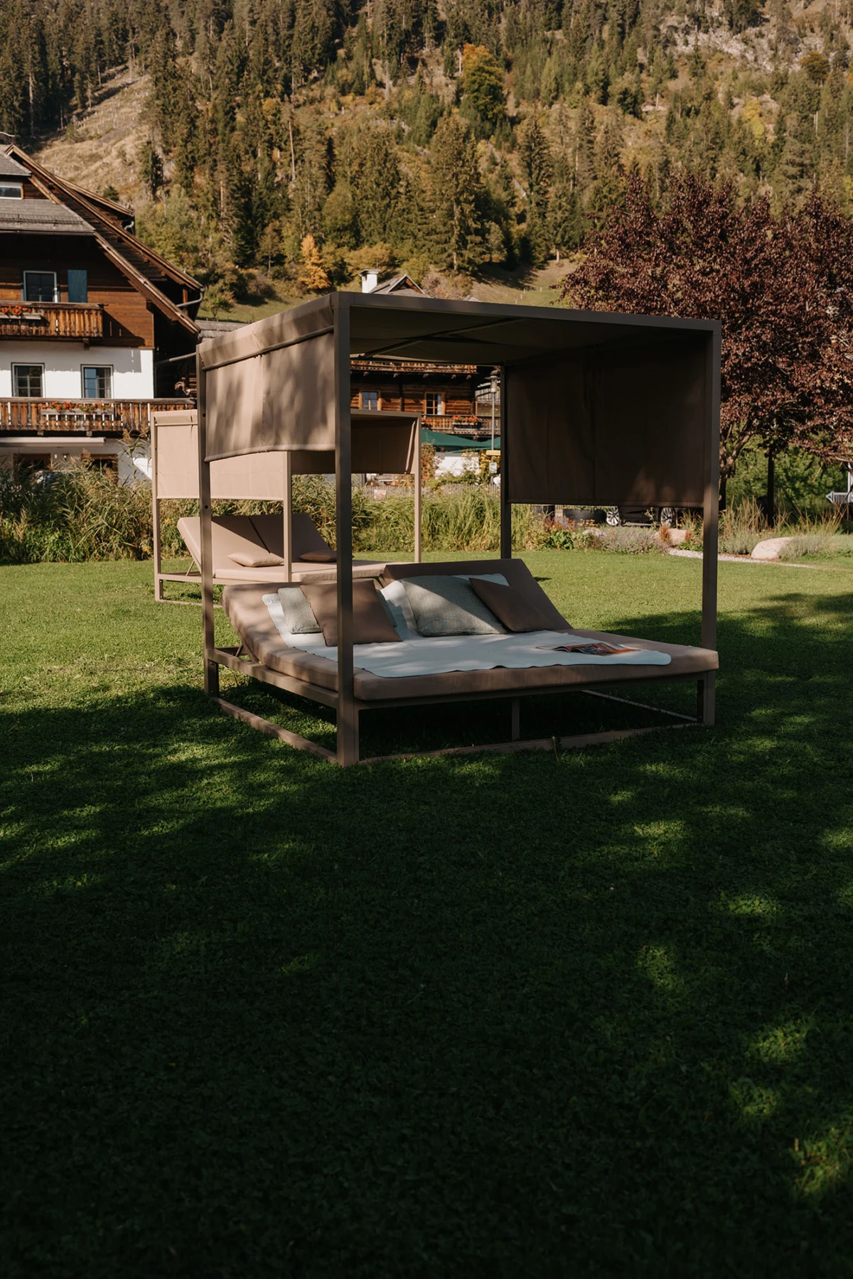 Freistehendes Outdoor-Liegebett mit Stoffdach und Kissen auf grünem Rasen; im Hintergrund Chalethäuser und Bergwald.