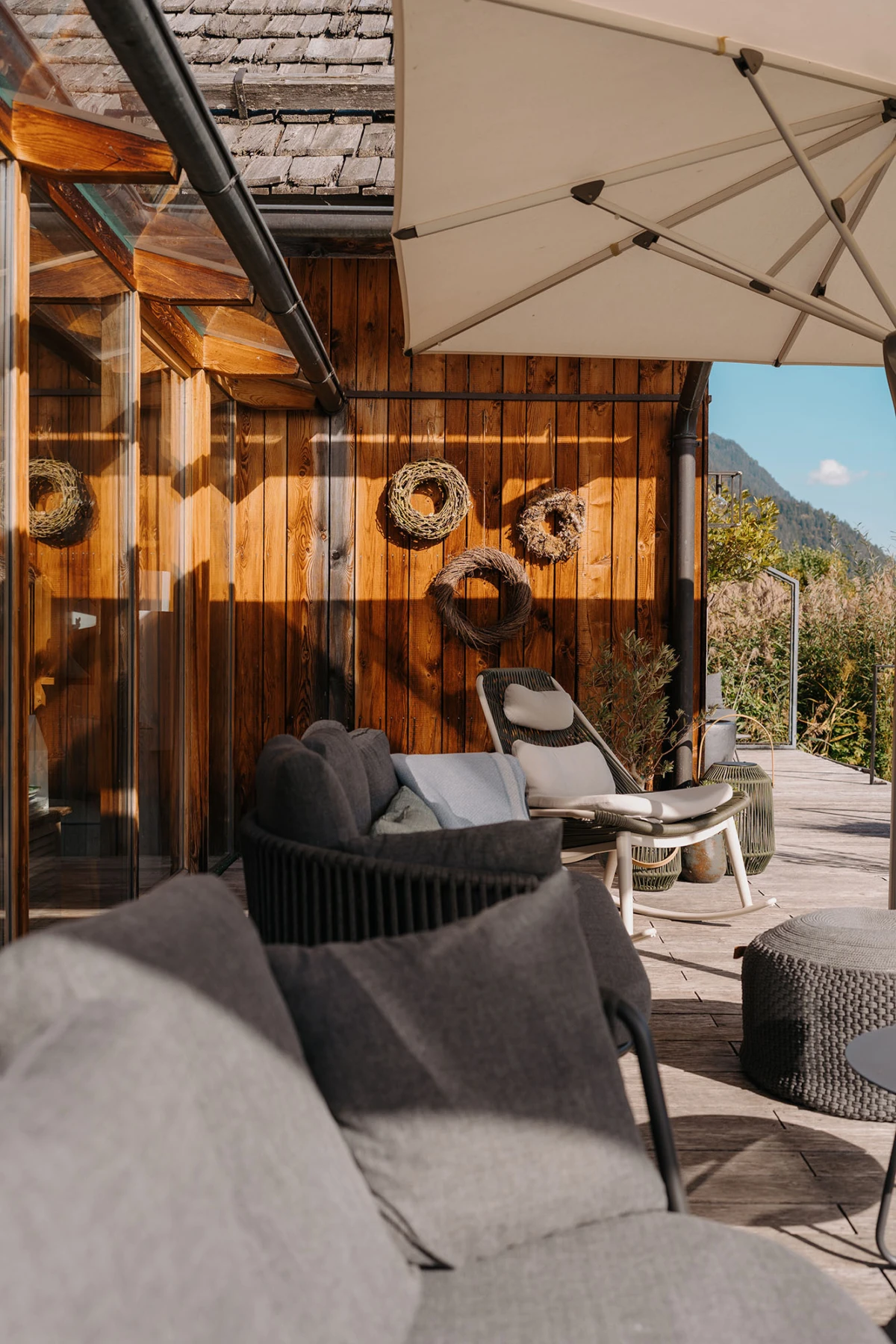 Outdoor-Terrasse mit grauem Polstersofa, Lounge-Sessel, großem Sonnenschirm; Holzwand mit dekorativen Zweigkränzen und Bergblick.