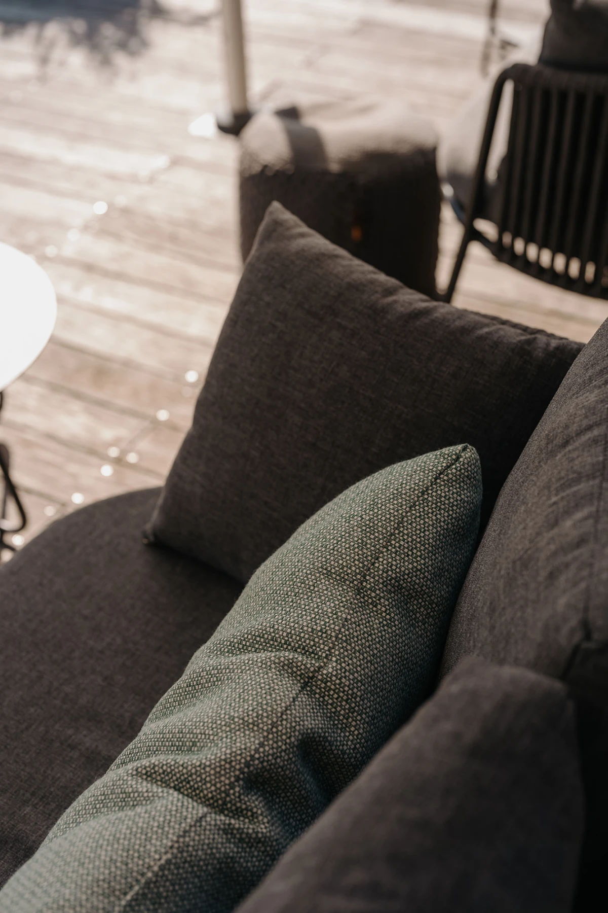 Außen-Sofa mit dunklem Bezug und grün-gemustertem Kissen im Vordergrund, Holzdeck und weitere Gartenmöbel im Hintergrund.