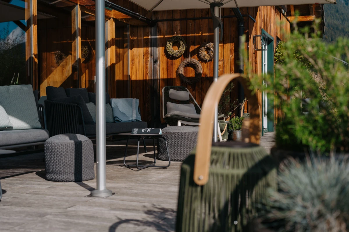 Gemütliche Outdoor-Lounge auf Holzveranda; Sofa, Korb-Sessel, Beistelltisch, Wandkränze, Pflanzen im Vordergrund.