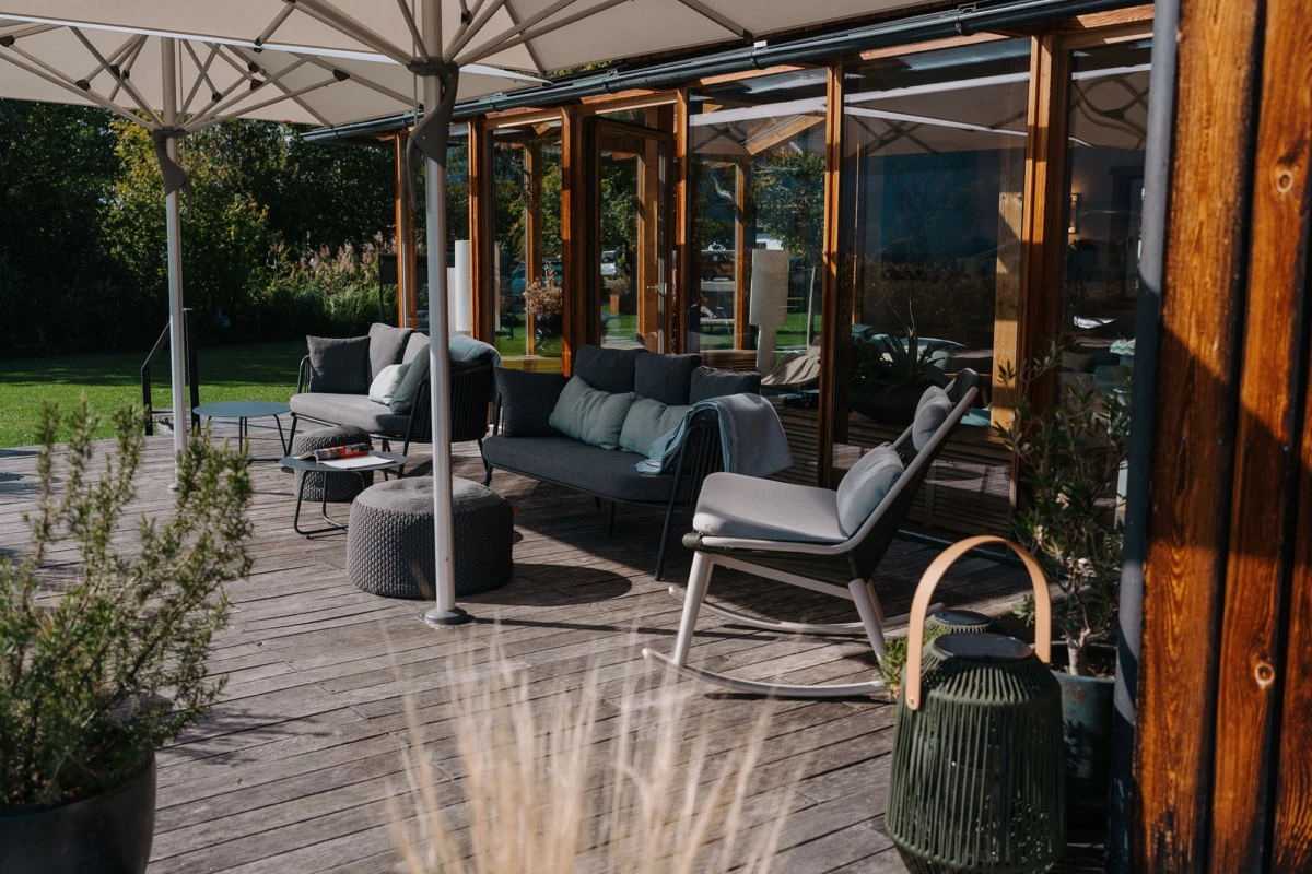 Holzterrasse mit grauen Lounge-Möbeln, Sofa, Sessel und Ottomane unter Sonnenschutz; Glas-Wand mit Holzrahmen, Garten im Hintergrund.