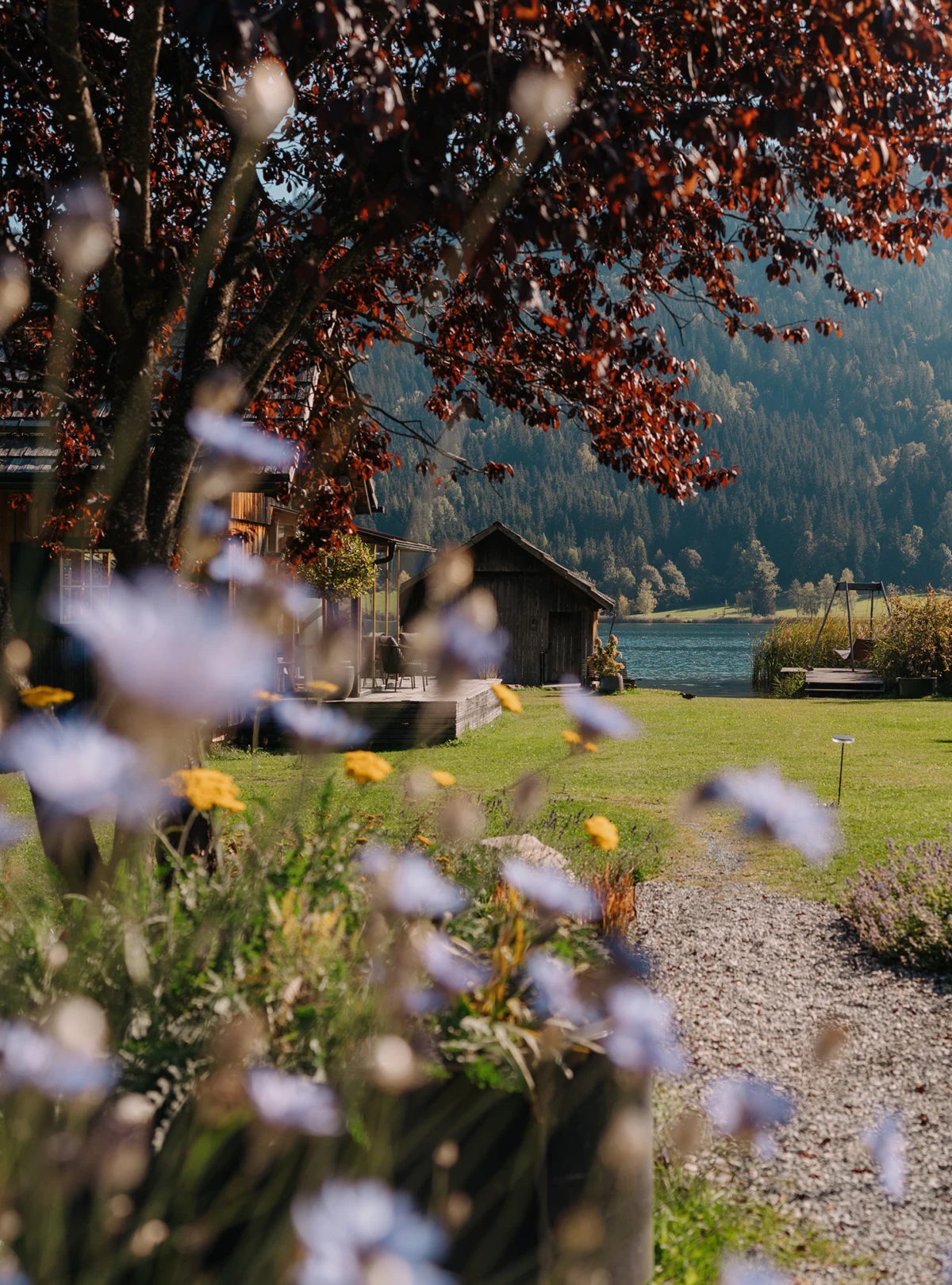 Holzhaus am Seeufer, Garten mit bunten Blumen im Vordergrund; Herbstlaub an Bäumen, Berglandschaft im Hintergrund.