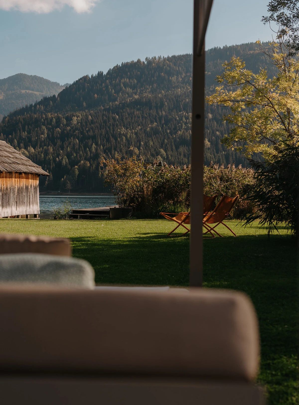 Seeuferlandschaft mit Holzhaus, grüne Wiese und zwei orangefarbene Liegestühle; dahinter Wald und Bergkette, Blick von einer Terrasse.