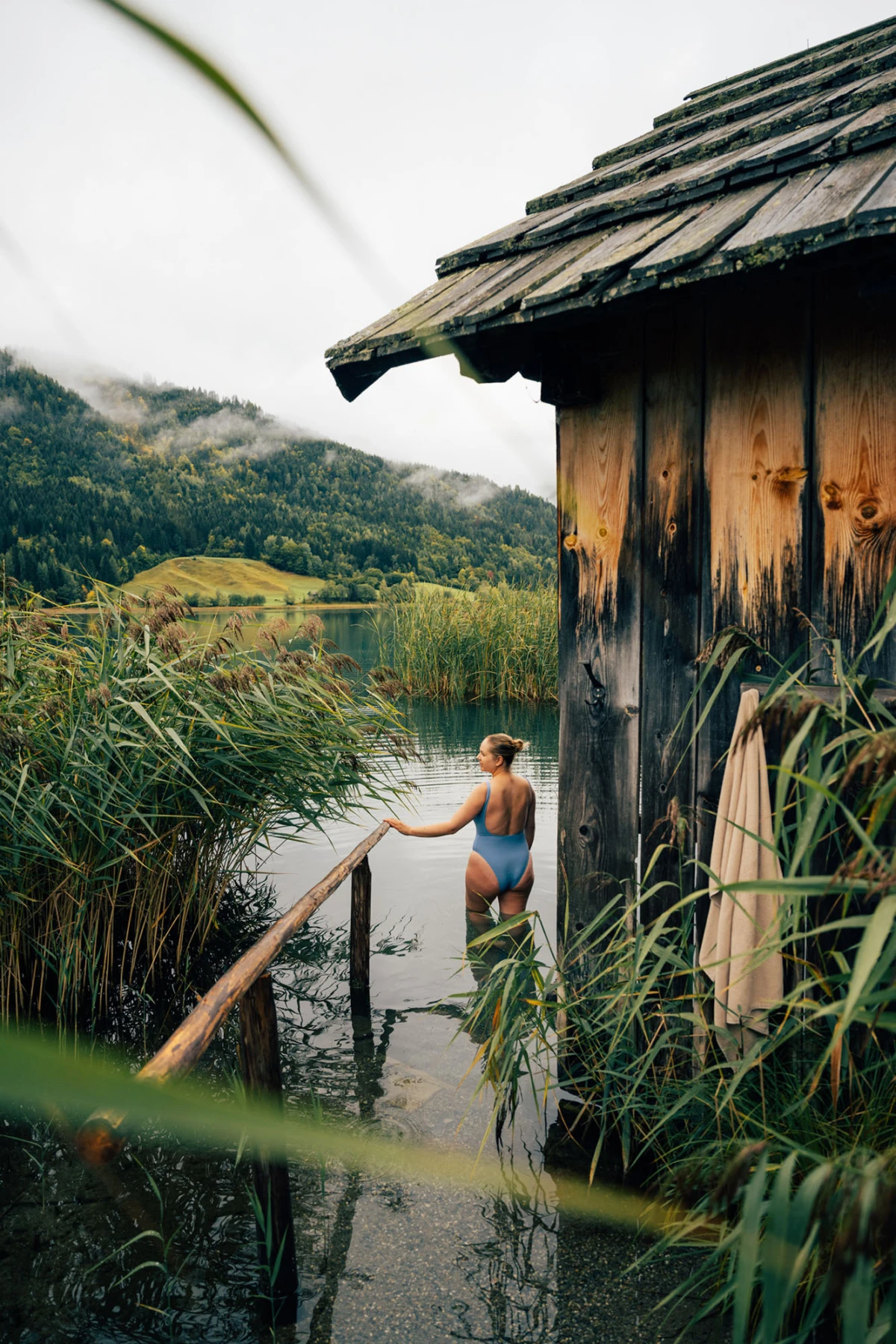 Person im blauen Badeanzug steht am Holzsteg neben einem rustikalen Seehaus, von Schilf umgeben, Blick auf See und Berge.