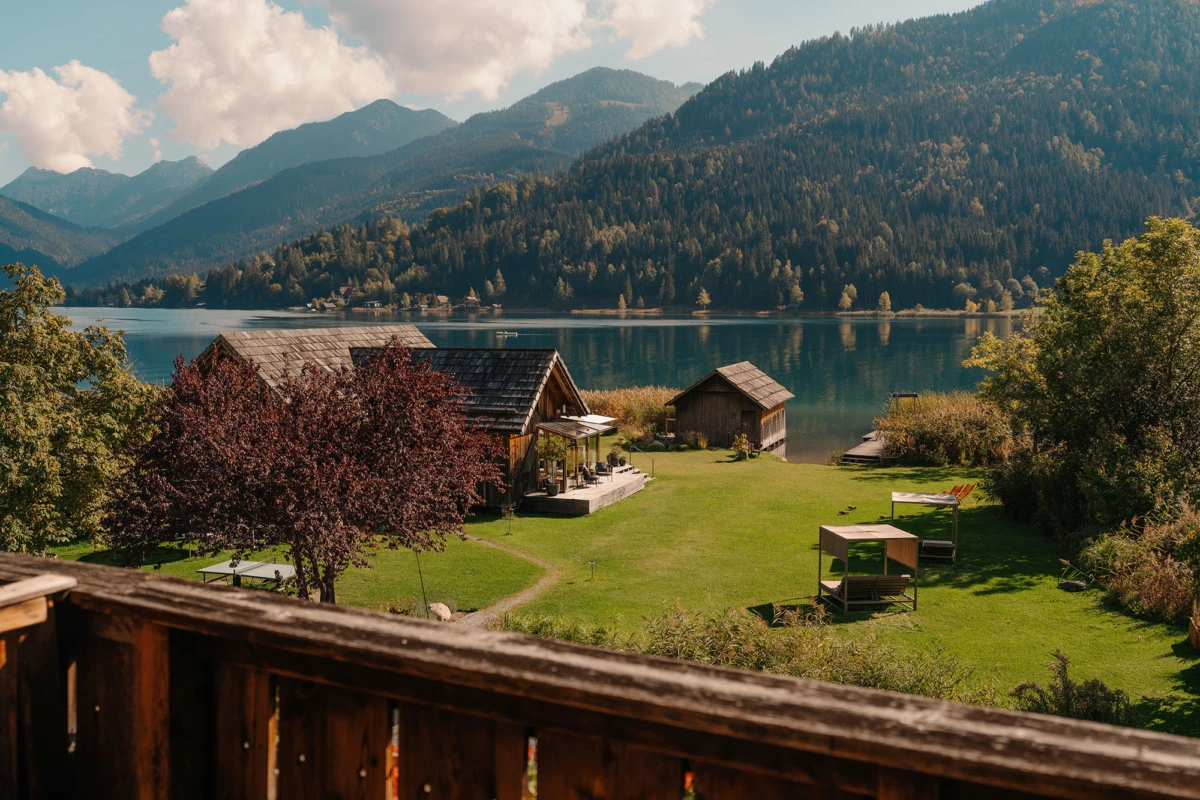 Seeufer mit Holzhäusern, Gartenmöbeln und Bäumen, grüne Wiese, Blick auf den See und bewaldete Berge im Hintergrund.