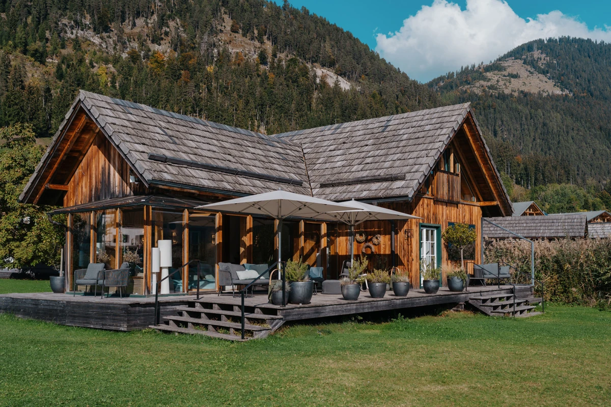 Holz-Chalet mit Veranda, Loungemöbel und Sonnenschirm, Topfpflanzen, vor Bergwaldlandschaft.