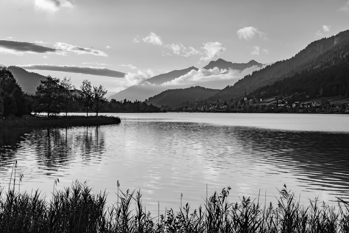 Schwarzweiß-Bergsee mit Ufergras im Vordergrund, ruhige Wasserfläche spiegelt Wolken und Berge; Dorf am Ufer in Fernsicht.
