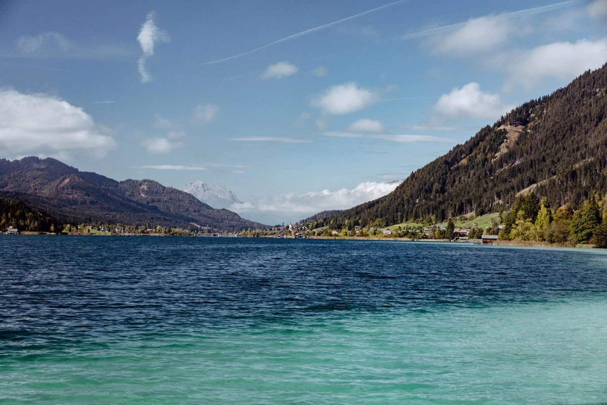 Bergsee mit türkisblauem Wasser, von Waldbergen umrahmt; Dorf am Ufer mit Häusern, blauer Himmel und Wolken.