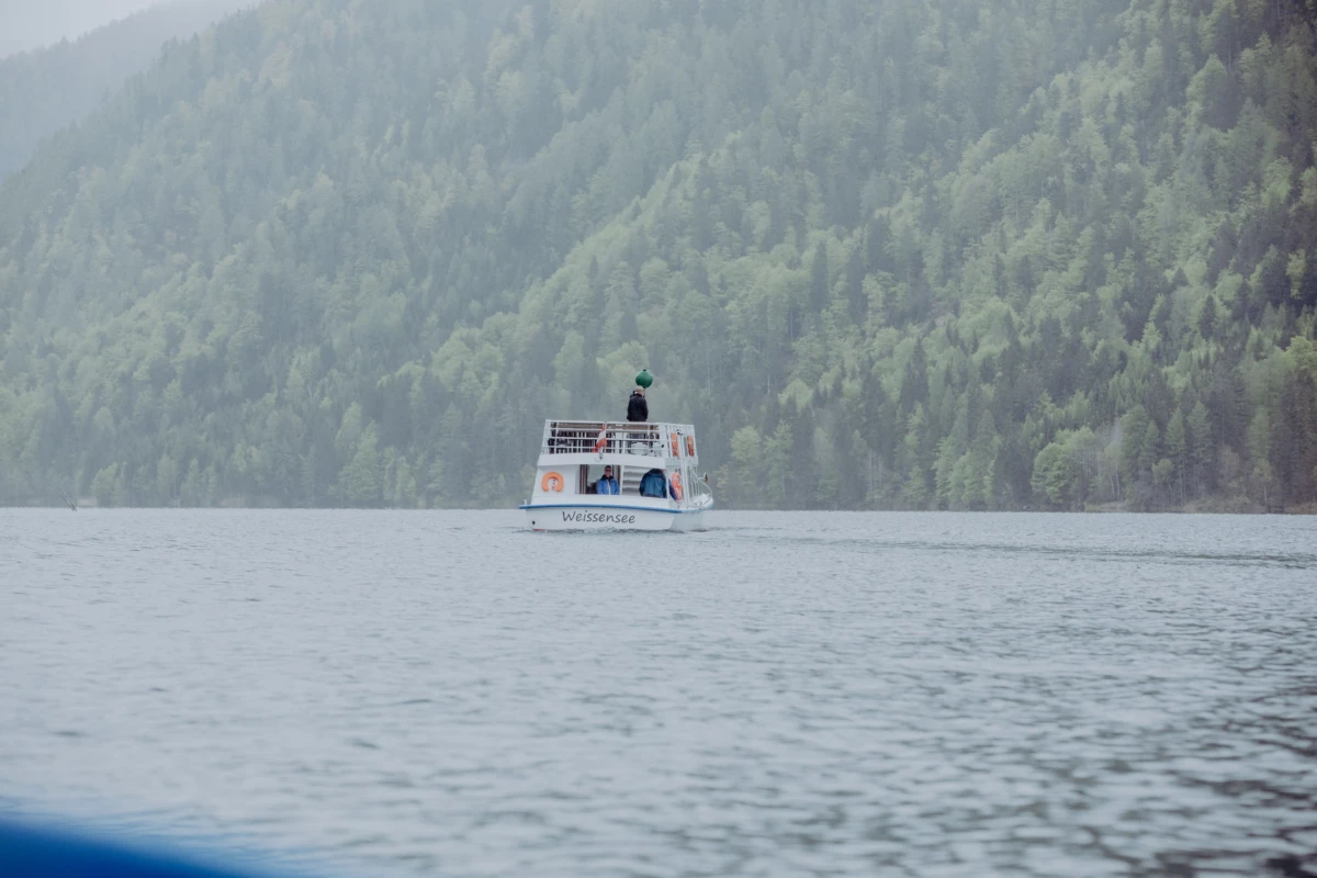 Passagierboot fährt über den Weissensee; Person oben auf dem Oberdeck; Waldgebirge im Hintergrund.