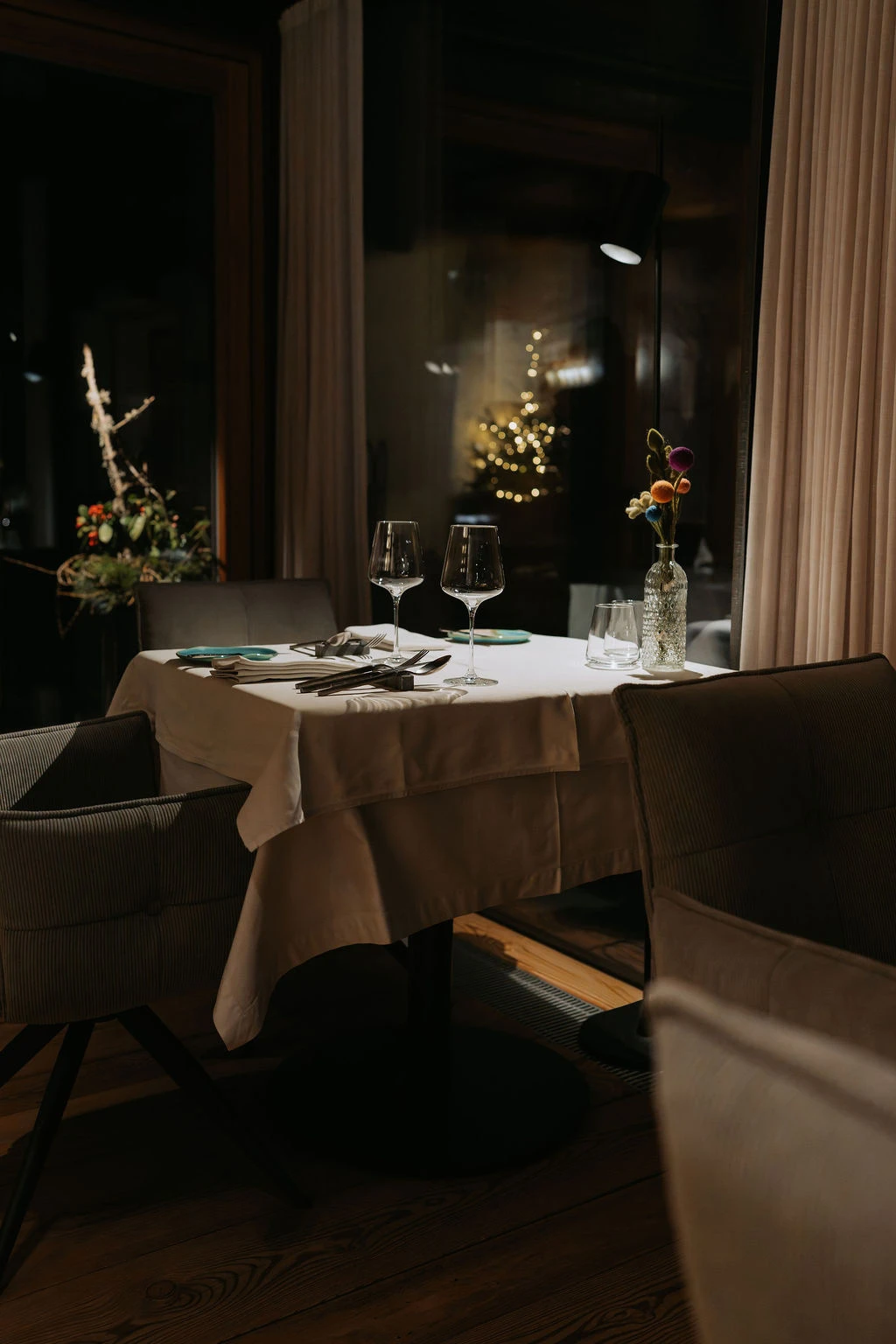 Gedeckter runder Tisch im eleganten Restaurant; zwei Weingläser, Wasserglas, Besteck, Blumenvase; warme Beleuchtung, Vorhänge.