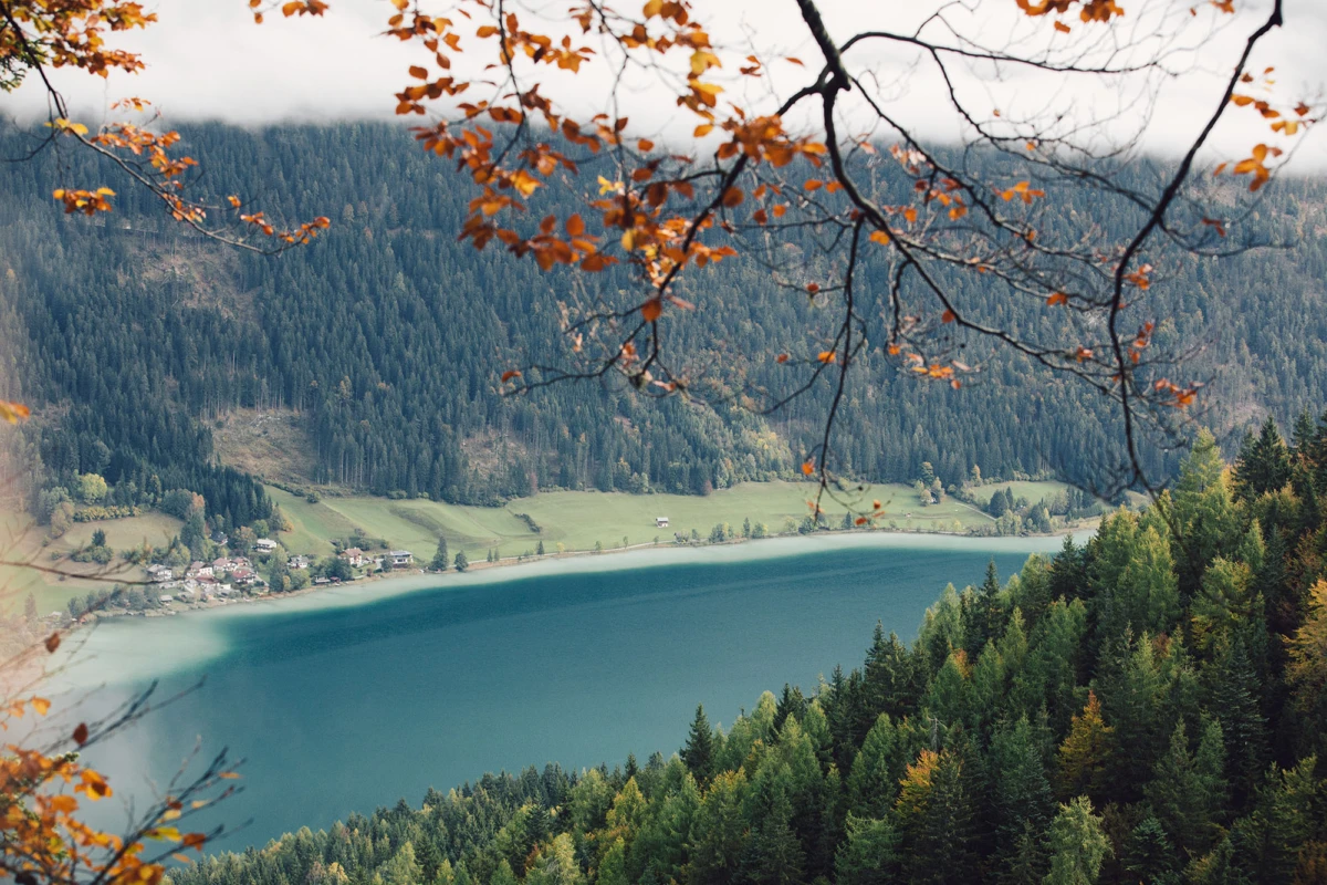 Türkisfarbener Bergsee, von Wald umgeben; Herbstlaub im Vordergrund, Dorf am Ufer, bergige Landschaft dahinter.